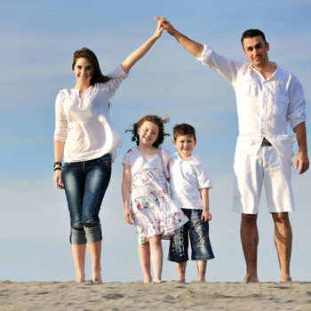 Family of four holding hands on the beach under a clear sky.