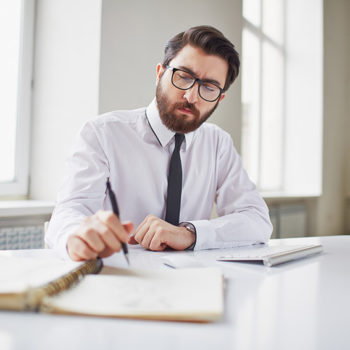 A focused man in glasses works at a desk with documents and a calculator.