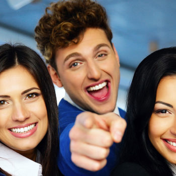 Three cheerful young adults smiling, with one pointing at the camera.